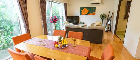 Un coin repas avec une table en bois, des chaises orange et un saladier rempli de fruits. On aperçoit un canapé et un téléviseur au fond.