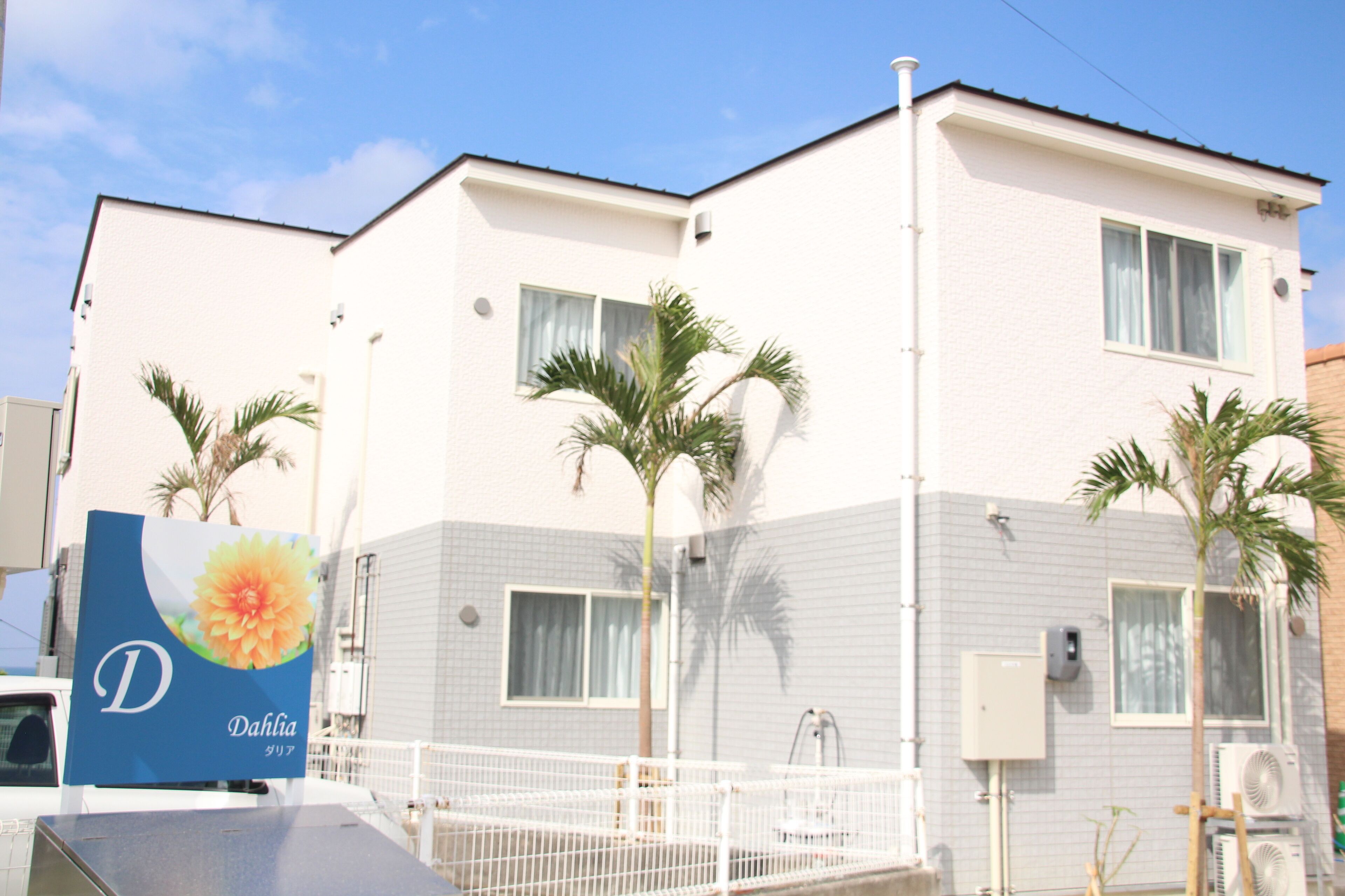 A two-story building with a white exterior, multiple windows, and a fenced outdoor area with palm trees.