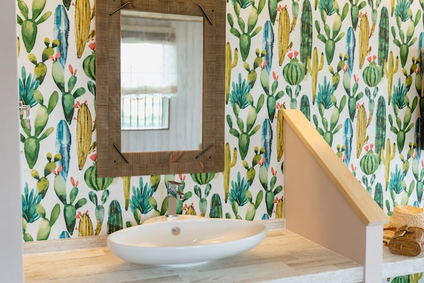 Un baño con lavamanos blanco, encimera de madera y un espejo encima. La pared está decorada con un papel tapiz estampado con motivos de cactus.