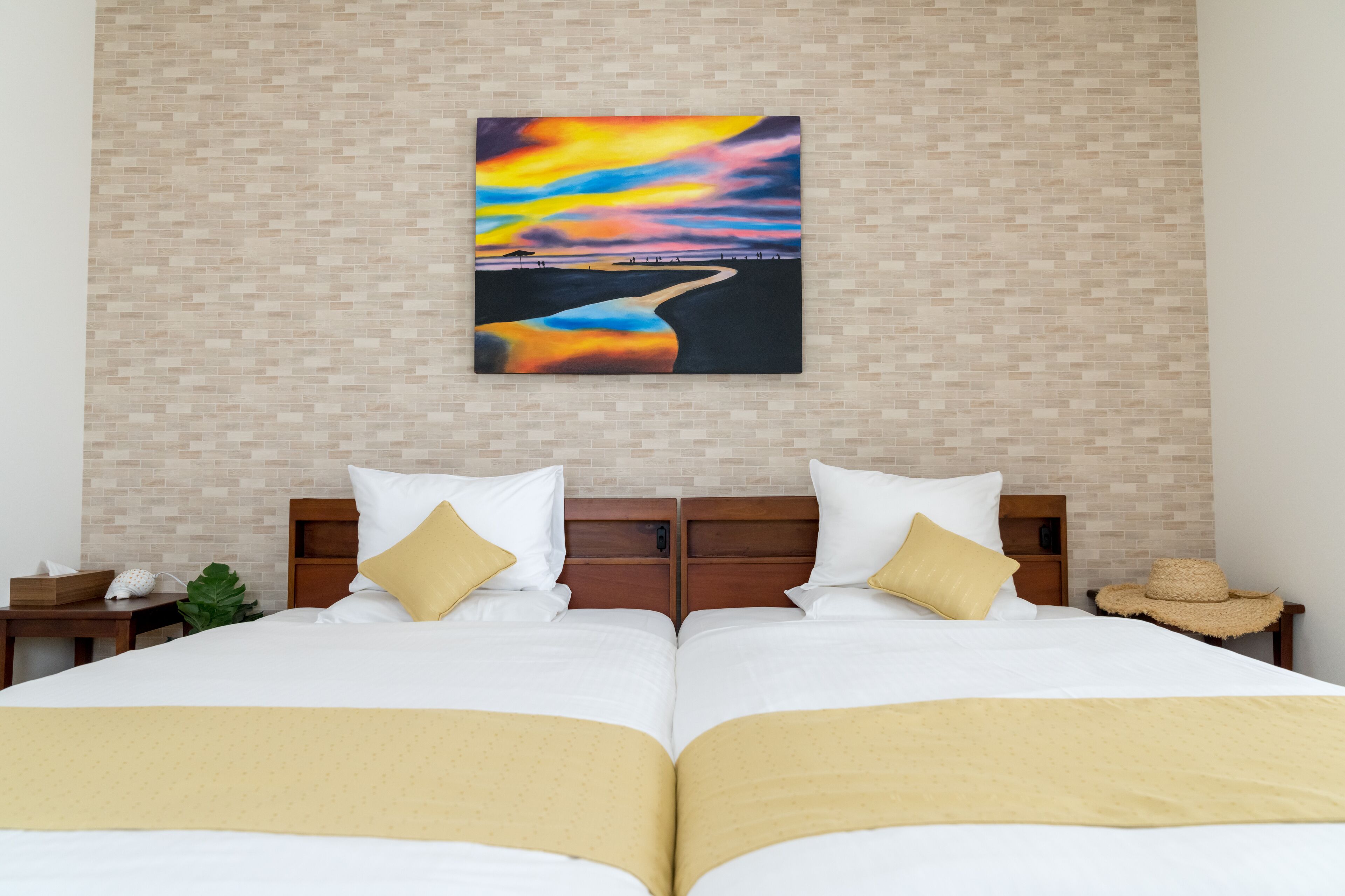 A double bed with white and gold bedding, a wooden headboard, and a wall-mounted painting of a sunset over a river.