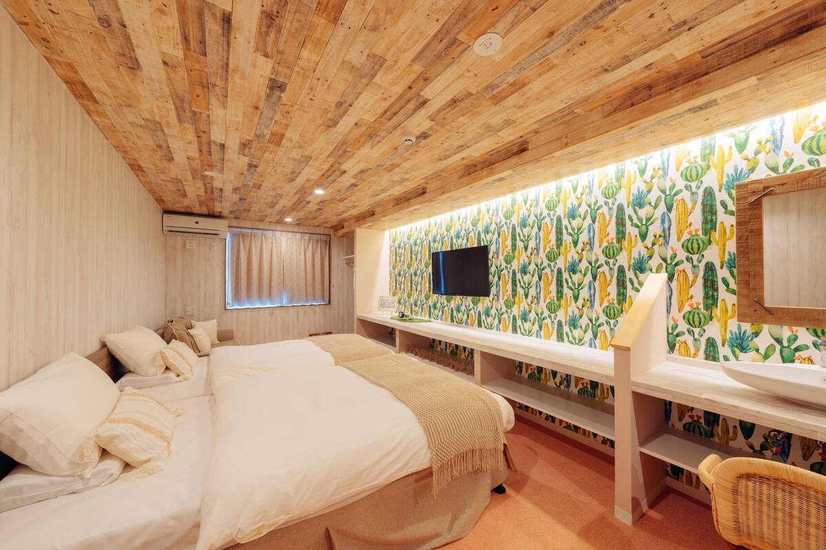Une chambre avec un plafond en bois, un lit avec une literie blanche, un téléviseur fixé au mur et un papier peint à motifs de cactus.