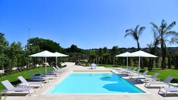 Una piscina con lettini bianchi e ombrelloni, circondata da vegetazione e palme.