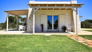 A covered outdoor seating area with stone walls and a wooden roof, adjacent to a building with white shutters and a glass door. - Borgo Fiorito Relais (Parabita)