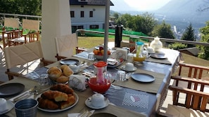 An outdoor dining setup with croissants, a fruit bowl, and a red drink on a table, overlooking a mountainous landscape. - B&B San Michele (Druogno)