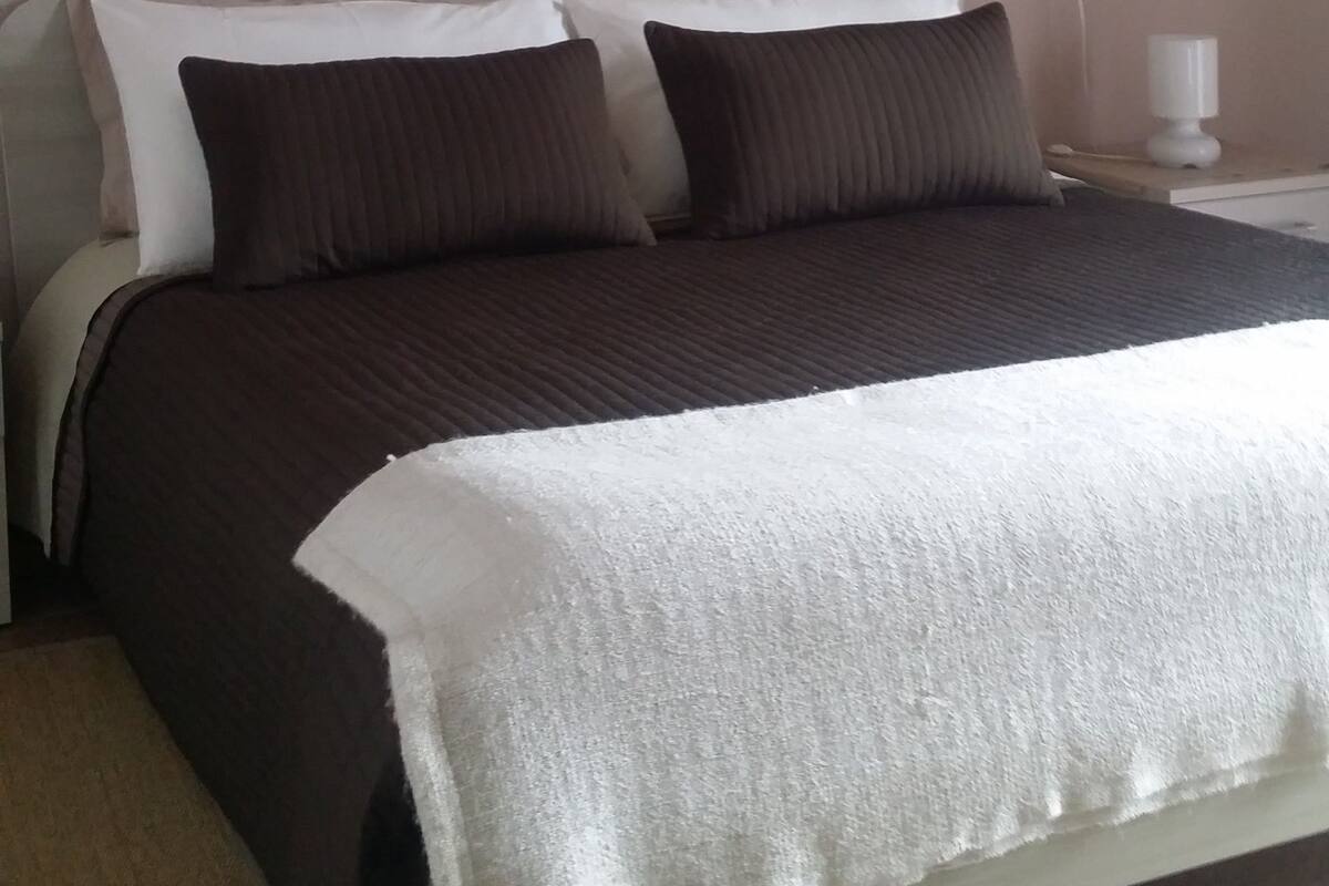 A bedroom with a bed, pillows, a bedside table with a lamp, and a painting of a pier on the wall.