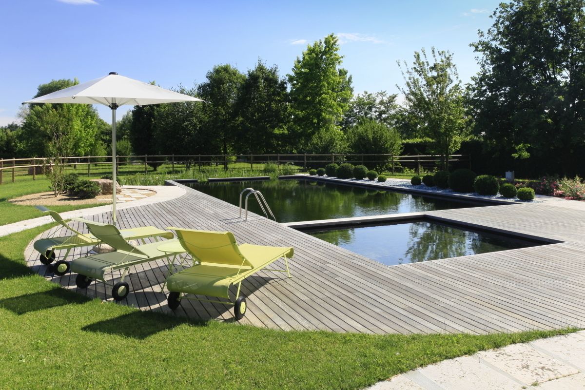 A deck with a pool, green lounge chairs, and a white umbrella.