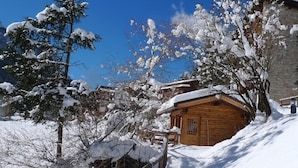 Une cabane en bois, dont le toit est recouvert de neige, entourée d’arbres et d’un mur de pierres.