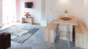 Un salon moderne avec un plafond en bois, un téléviseur à écran plat fixé au mur, une petite table en bois ornée d’un vase de fleurs et un banc avec une chaise en plastique transparent.