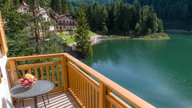 Un balcon avec une table et des chaises donnant sur un lac, entouré d’arbres et d’immeubles.