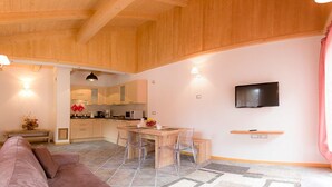 Un vaste espace de vie doté d’un plafond en bois, une cuisine à l’arrière-plan, une table à manger avec des chaises, un canapé et un téléviseur fixé au mur.