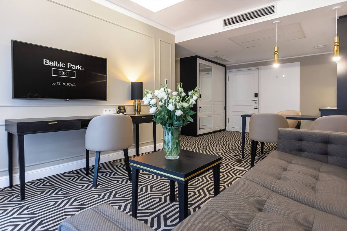 A modern hotel lobby with a large flat-screen TV displaying 'Baltic Park by ZDROJOWA', a black and white patterned floor, a small table with a vase of white flowers, and a grey sofa.