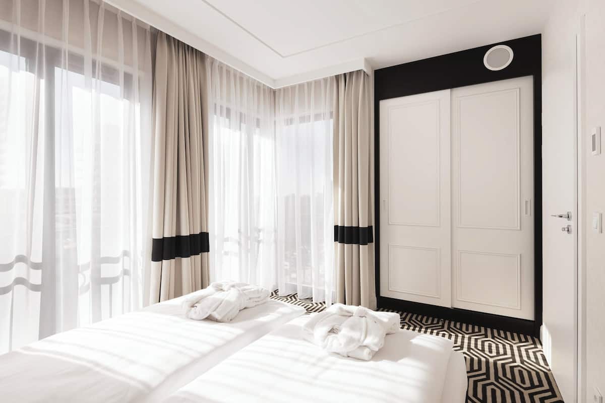 A hotel room with a large bed, sheer curtains, a black-framed wardrobe, and a patterned floor.