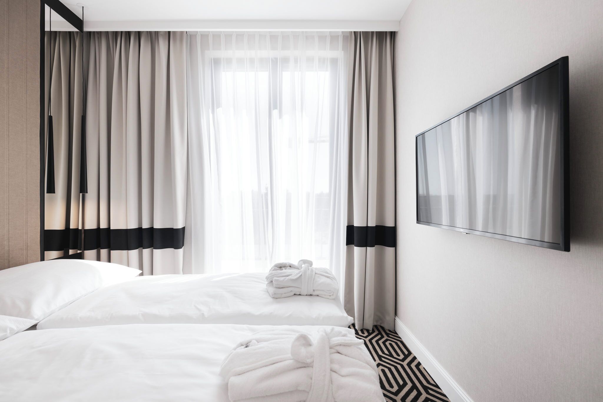 A hotel room with two beds, a flat-screen TV, and a window with sheer curtains.
