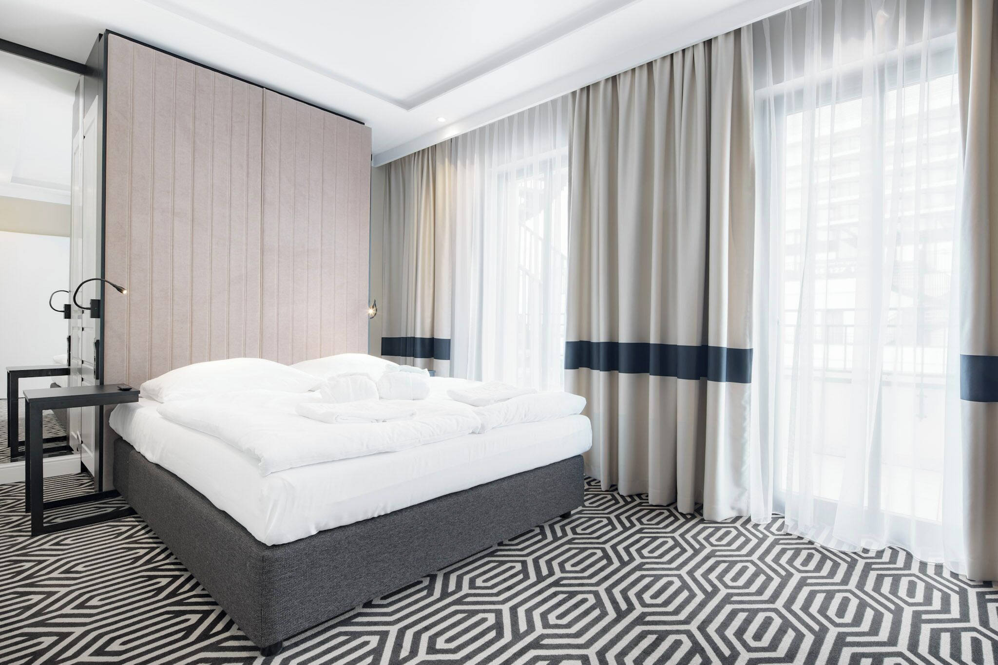 Quarto de hotel moderno com cama bem arrumada, mesa de cabeceira, janela grande com cortinas leves e tapete com padrão geométrico.