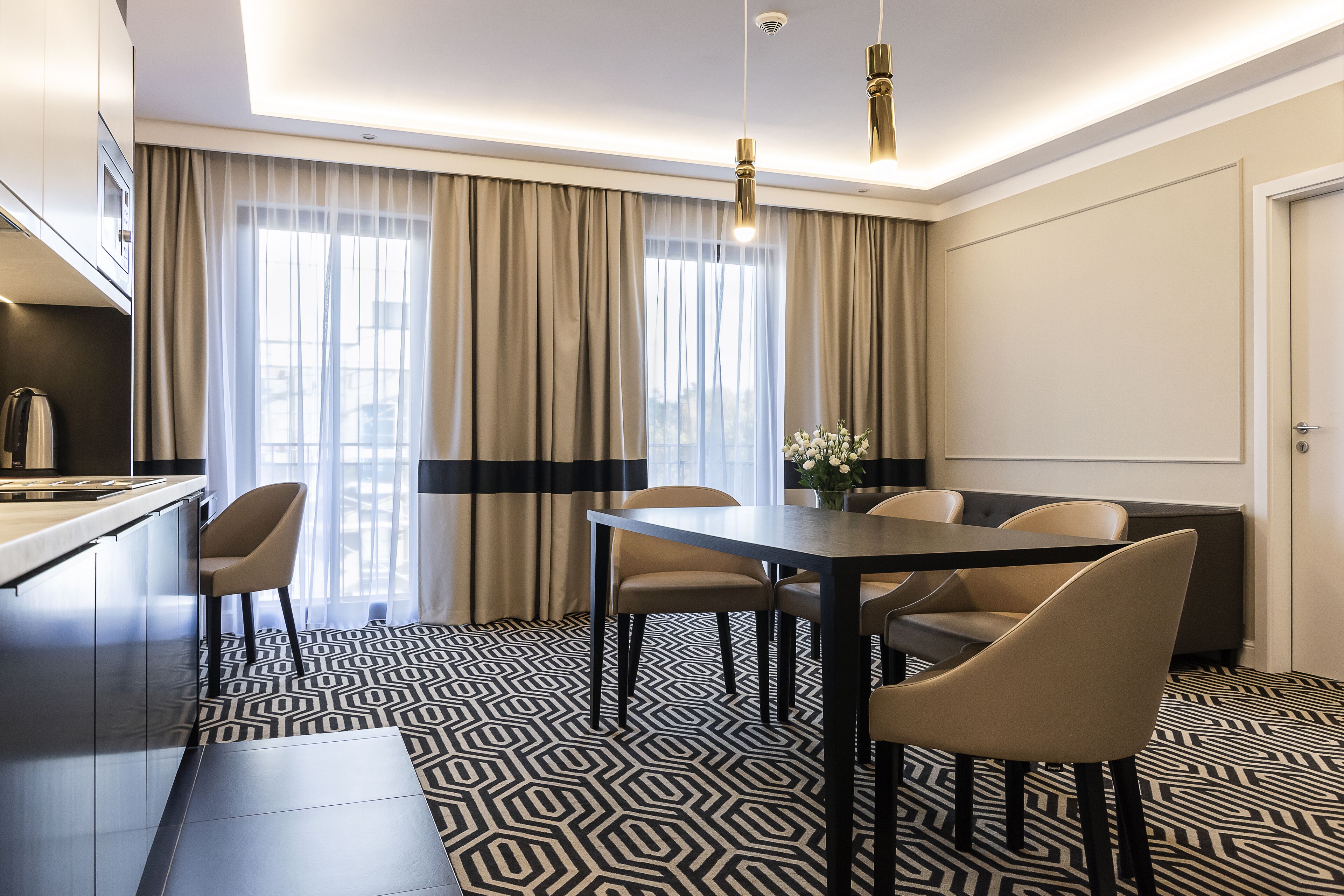 A modern hotel room with a dining area, a kitchen, and a large window with curtains.
