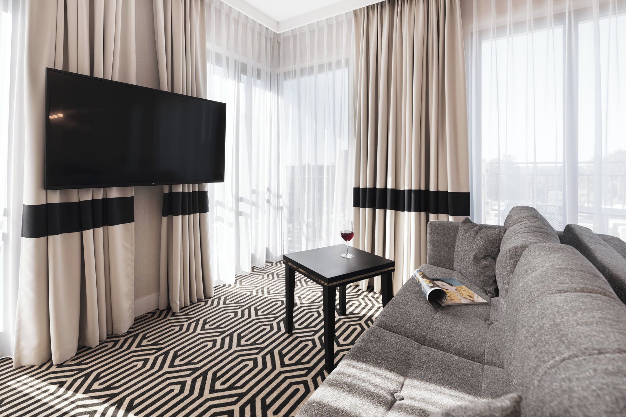 Ein modernes Hotelzimmer mit einem großen Flachbildfernseher an der Wand, einem grauen Sofa, einem kleinen schwarzen Tisch mit einem Glas Rotwein und einem gemusterten Boden.