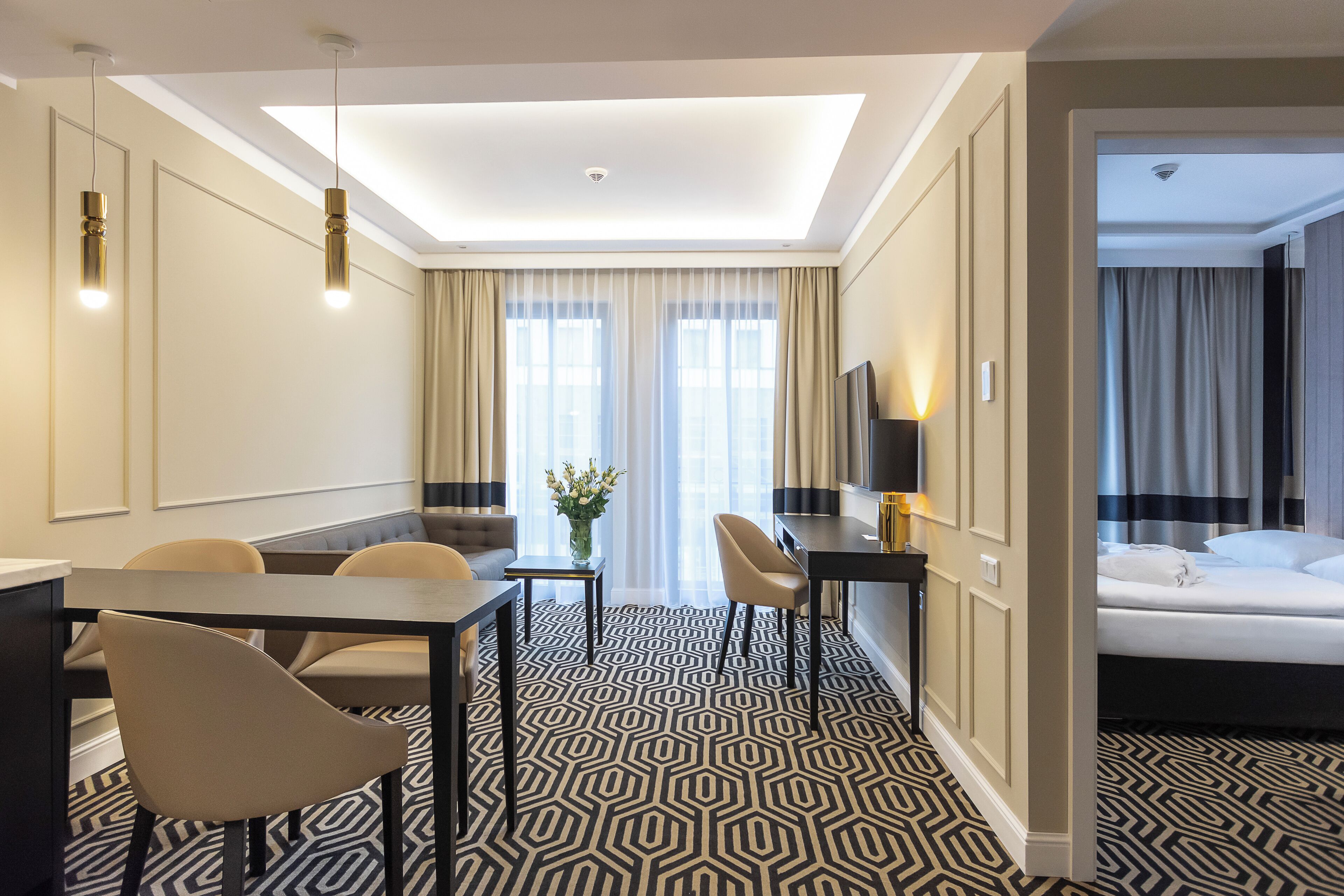 A hotel room with a modern design, featuring a large bed, a desk with a chair, a dining area with a table and chairs, and a patterned floor.