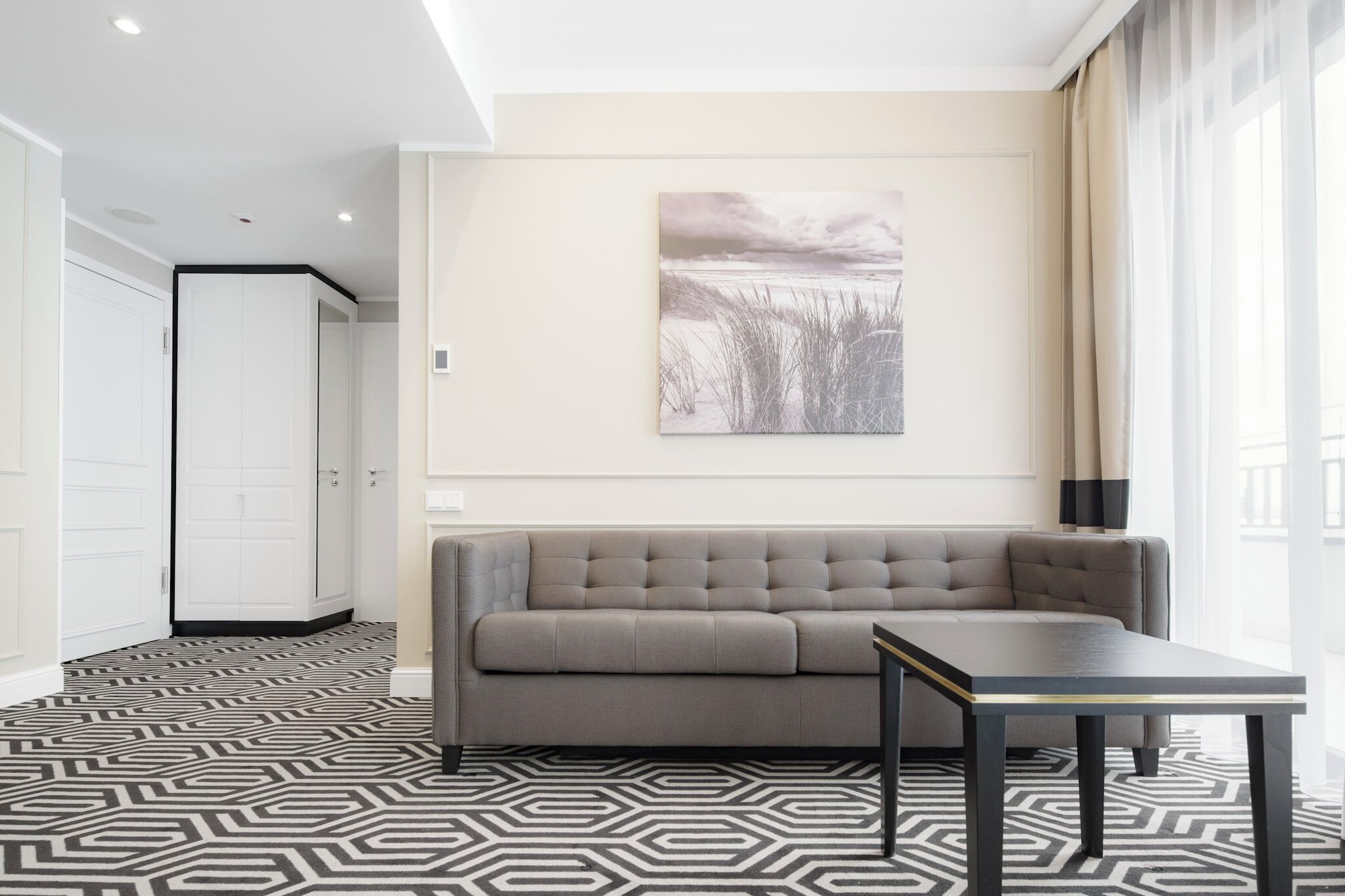 Une chambre d’hôtel moderne avec un canapé gris, une petite table, un tapis à motifs et un tableau représentant une scène de plage accroché au mur.