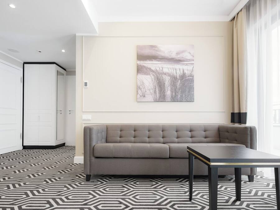 A modern hotel room with a grey sofa, a small table, a patterned rug, and a painting of a beach scene on the wall.
