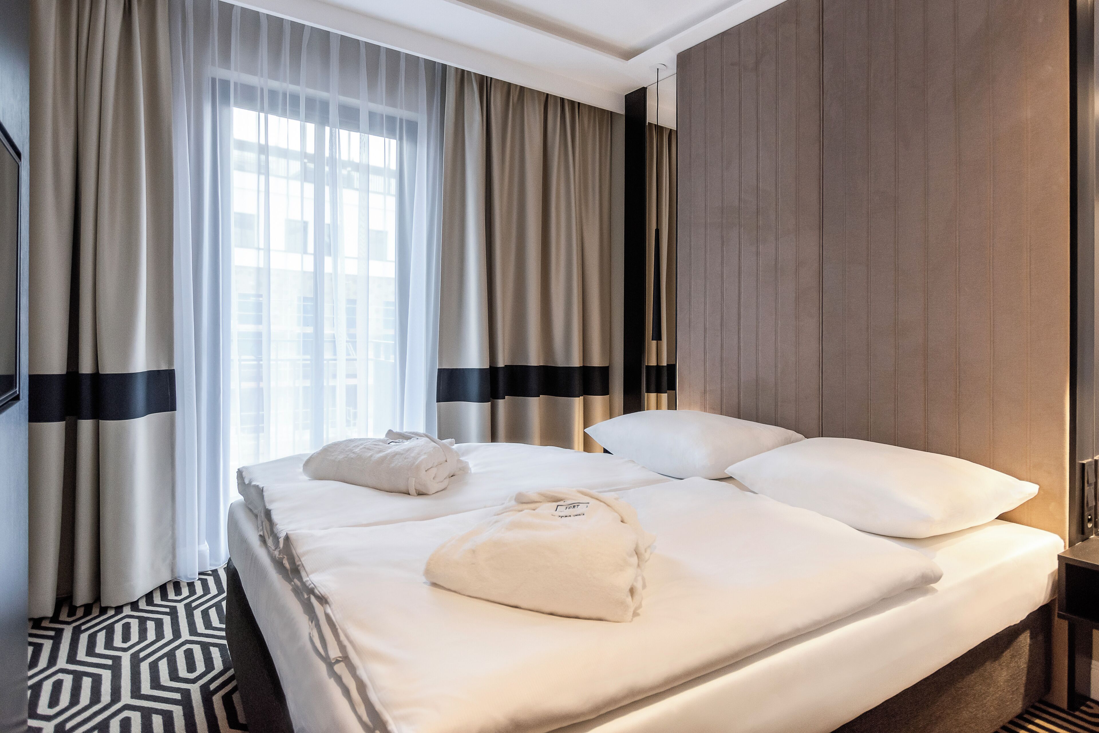 A hotel room with a neatly made bed, a large window with sheer curtains, and a geometric patterned floor.