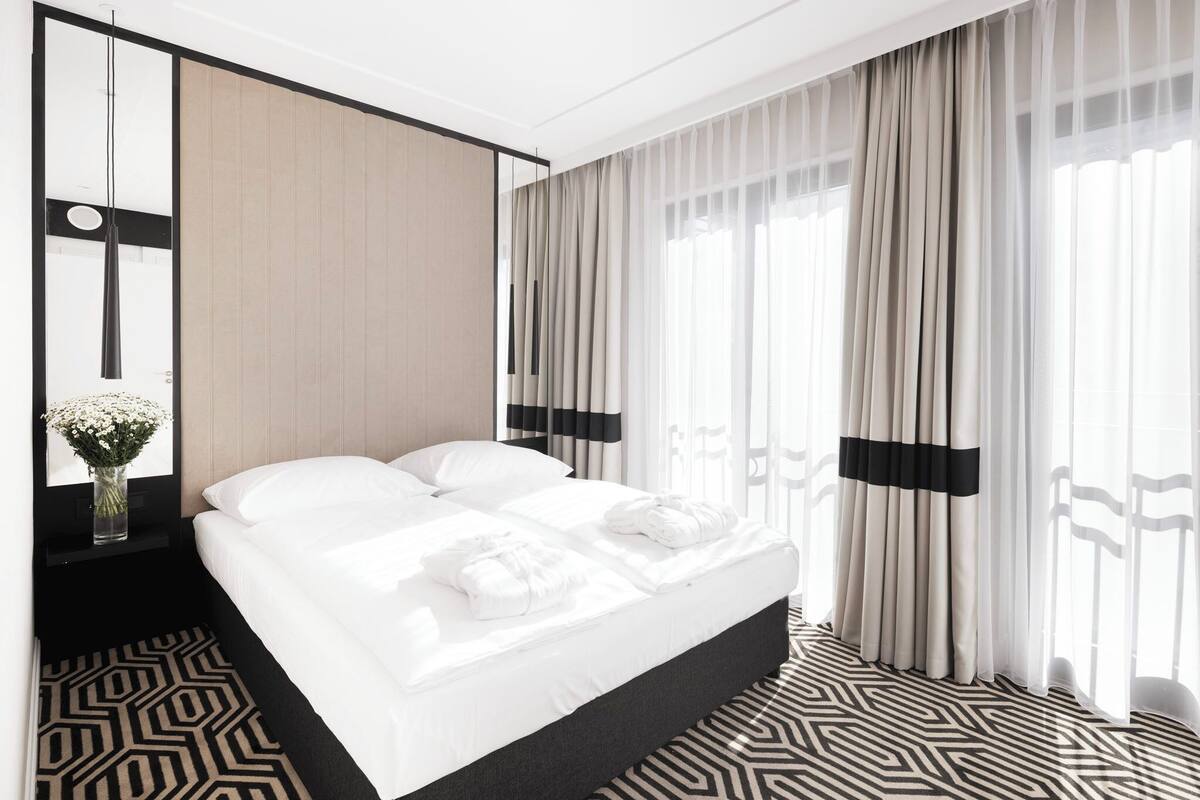 A modern hotel room with a large bed, a bedside table with a vase of flowers, sheer curtains, and a geometric patterned floor.