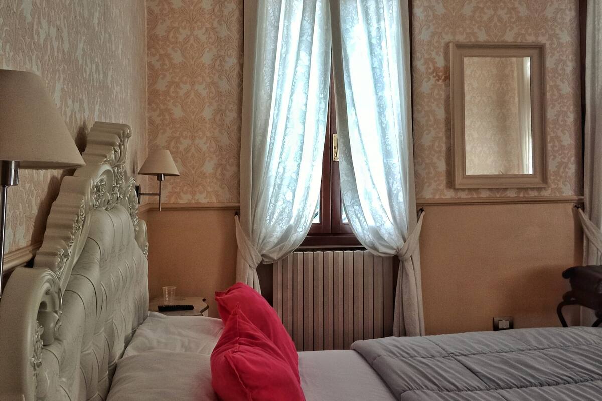 A hotel room with a large bed, a bedside table with a lamp, a radiator, and a window with sheer curtains.
