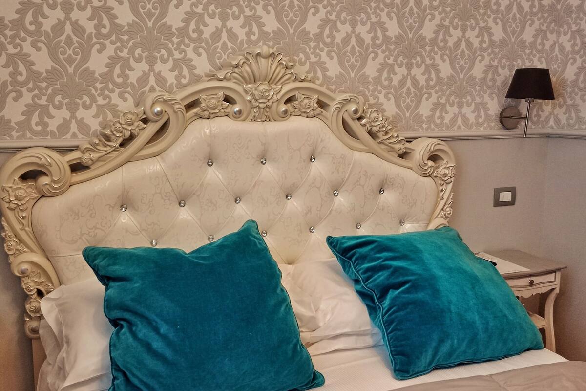 A bed with a tufted headboard, two teal pillows, and a white bedspread in a room with patterned wallpaper.