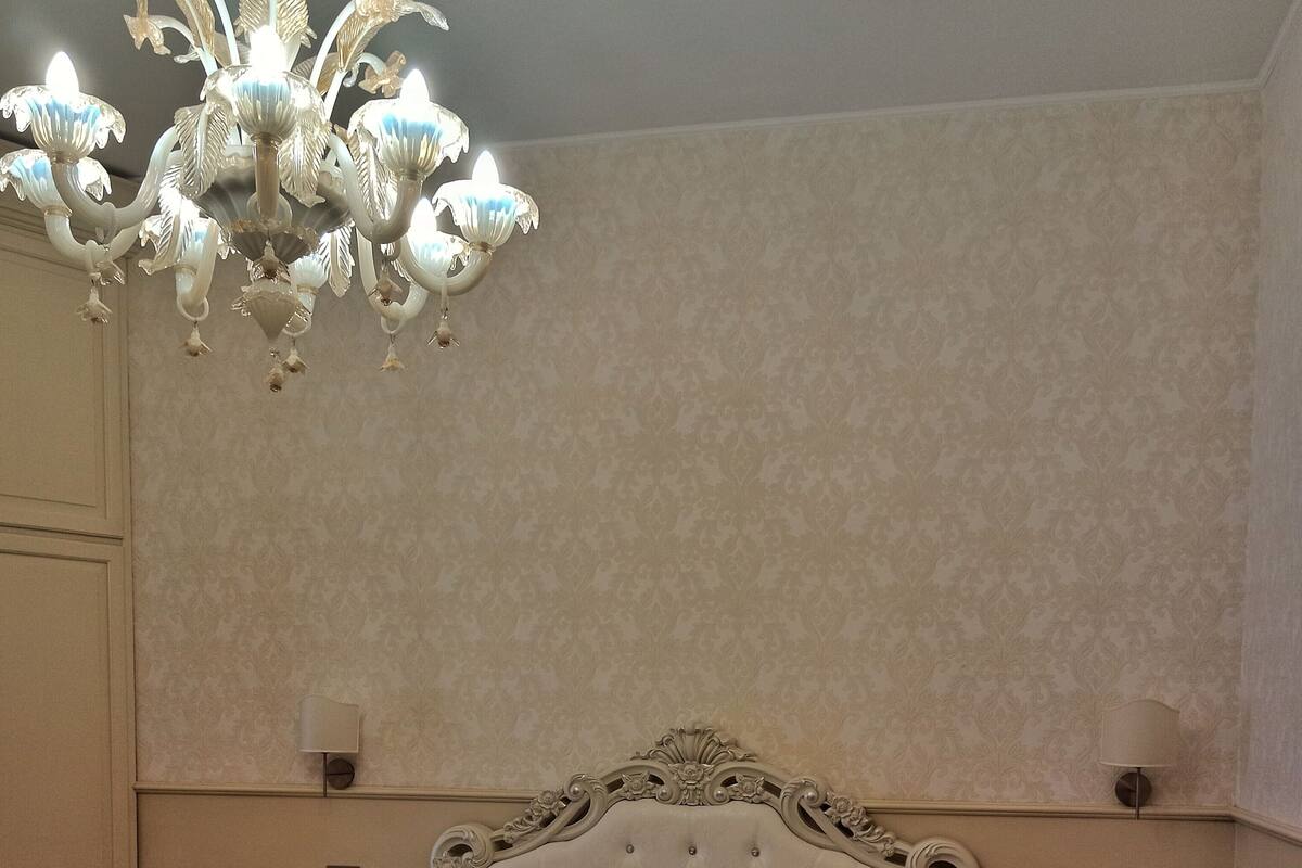 A bedroom with a tufted headboard, a chandelier, two bedside tables, and a patterned wall.