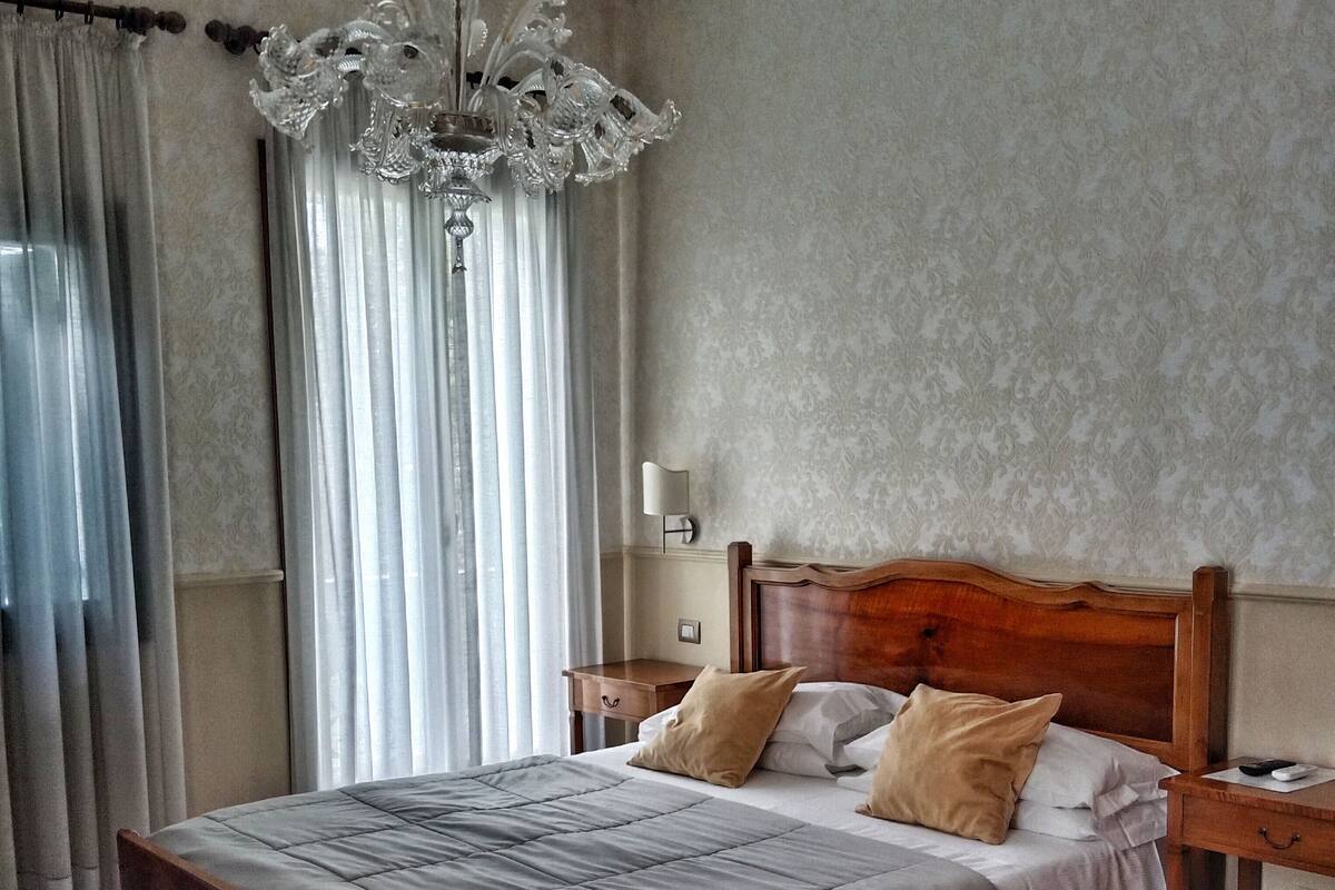 A bedroom with a wooden bed, a chandelier, a nightstand, and a wall-mounted lamp.