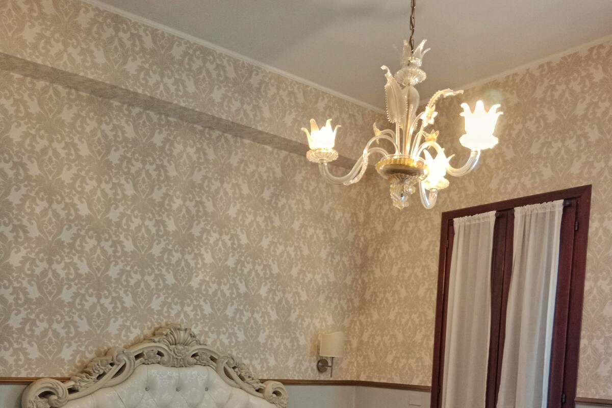 A bedroom with a tufted headboard, a chandelier, a bed with a grey bedspread and orange pillows, a nightstand, a wooden chair, and a window with curtains.