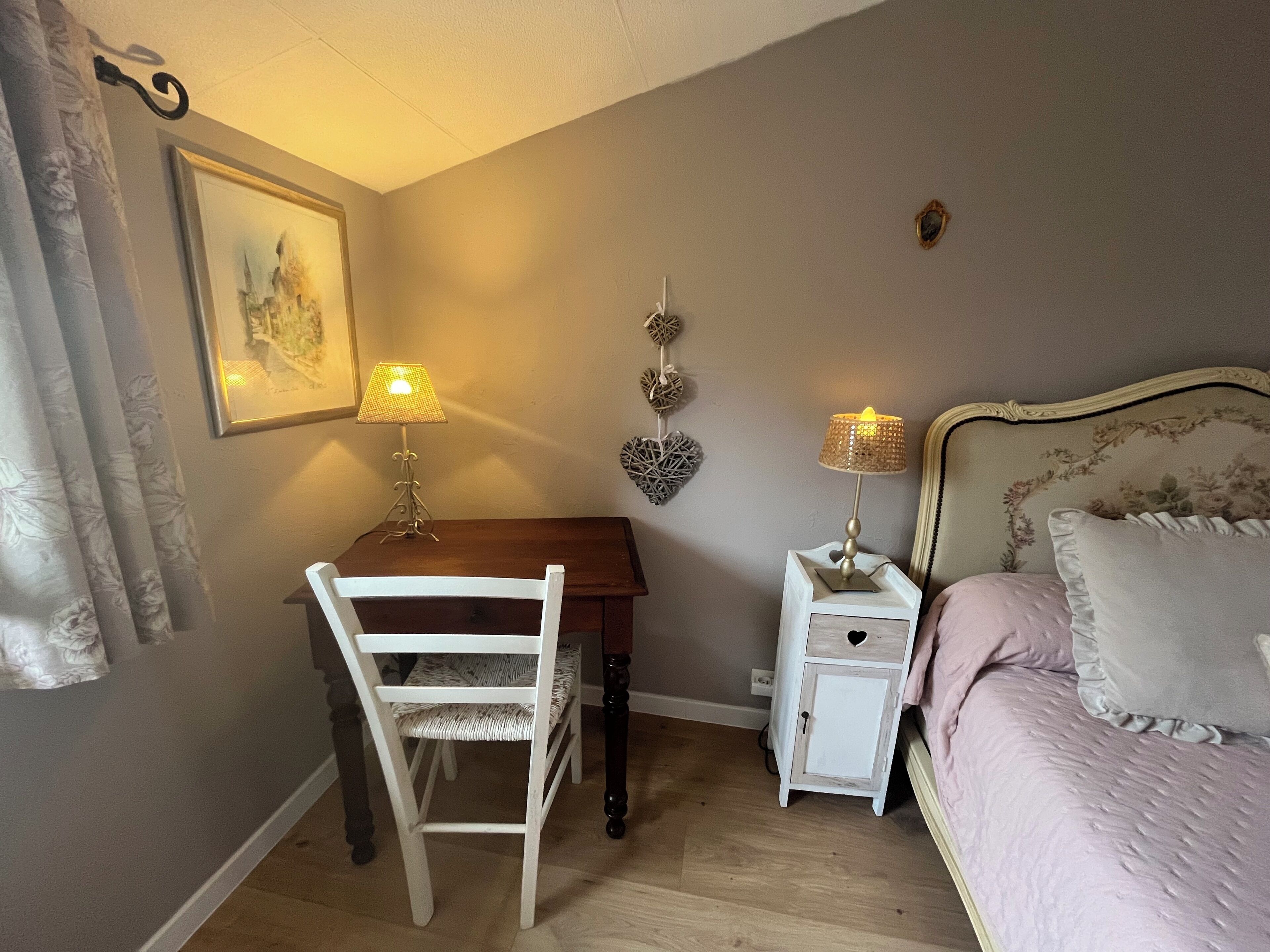Un dormitorio con un escritorio de madera, dos lámparas de noche, una cama con cabecera floral, una mesita de noche blanca y un cuadro colgado en la pared.