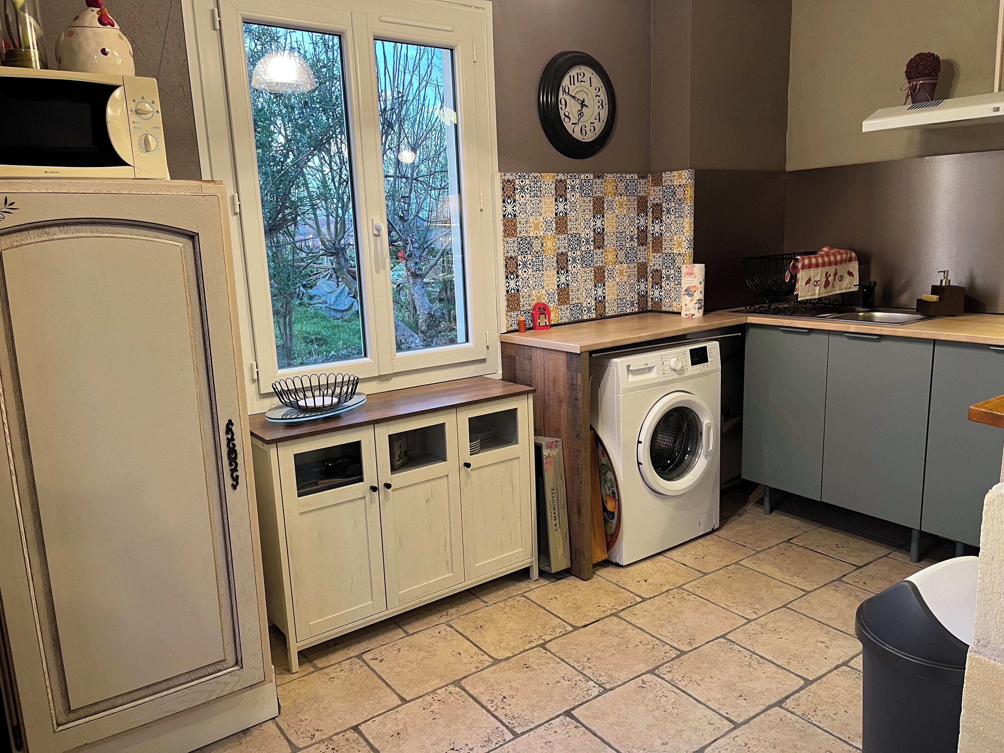 A compact kitchen with a washing machine, a microwave, a sink, and a window with a view of trees.