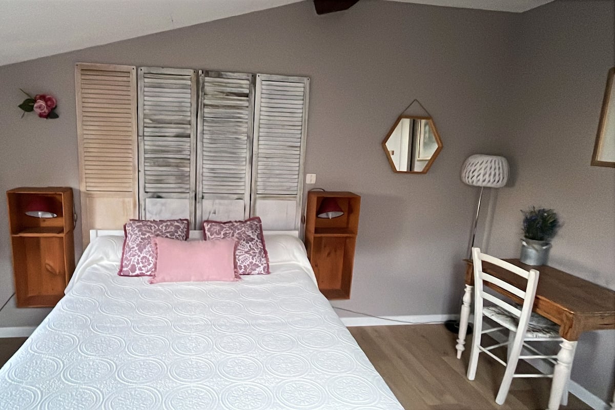 A bedroom with a bed, two wooden nightstands, a desk with a chair, a mirror, and a potted plant.