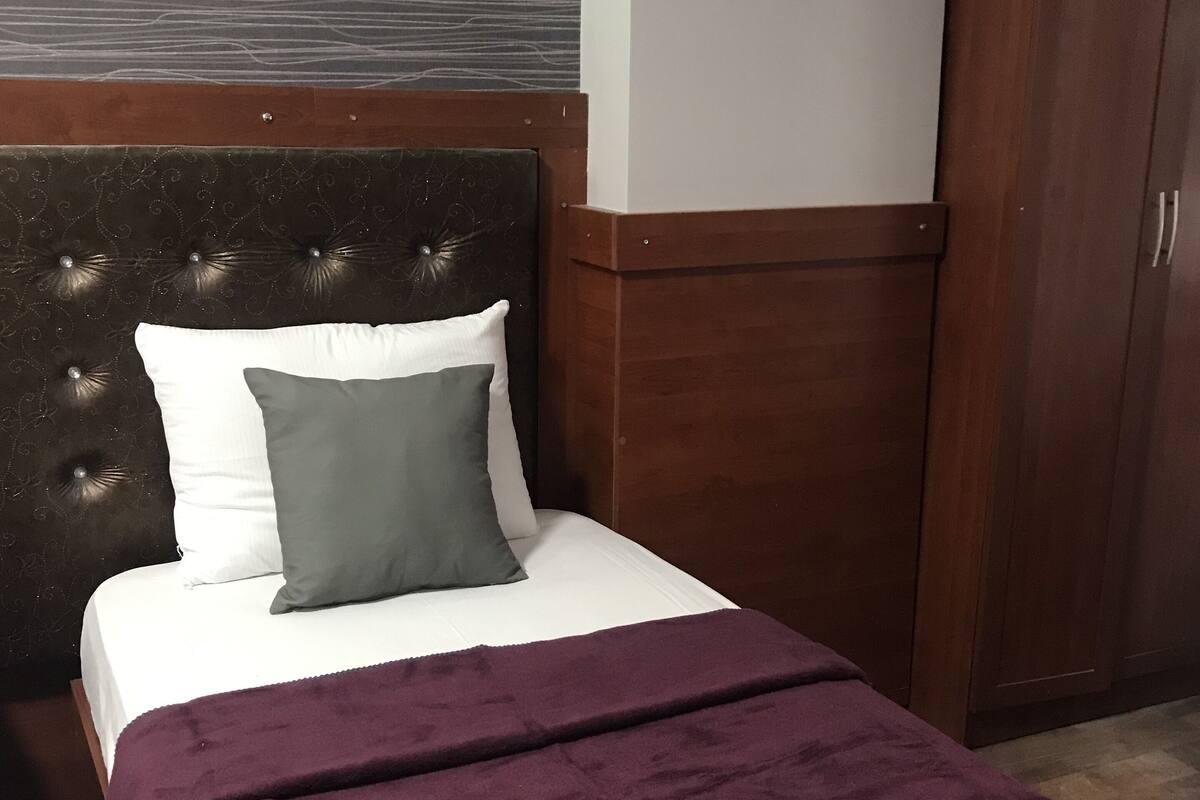 Un lit avec une tête de lit sombre, un oreiller blanc, un oreiller gris et une couverture violette.