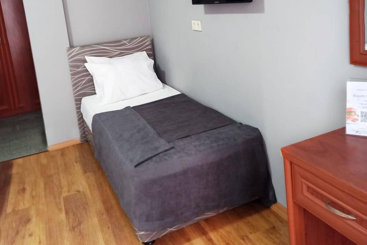 Une petite chambre avec un lit simple, une couverture grise, un téléviseur fixé au mur et un bureau en bois avec une chaise.