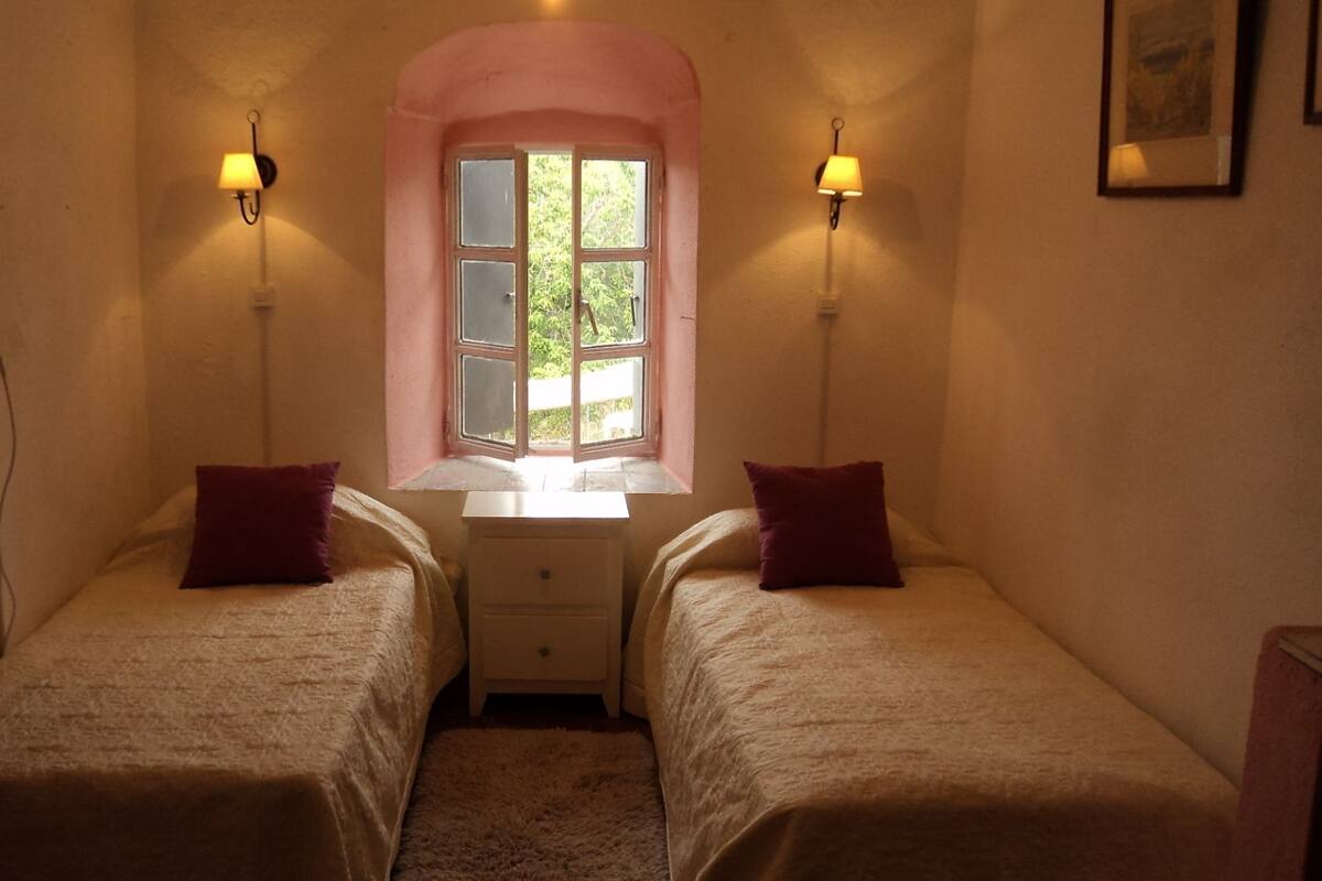 Une petite pièce avec deux lits, une fenêtre donnant sur un paysage verdoyant et des lampes fixées au mur.