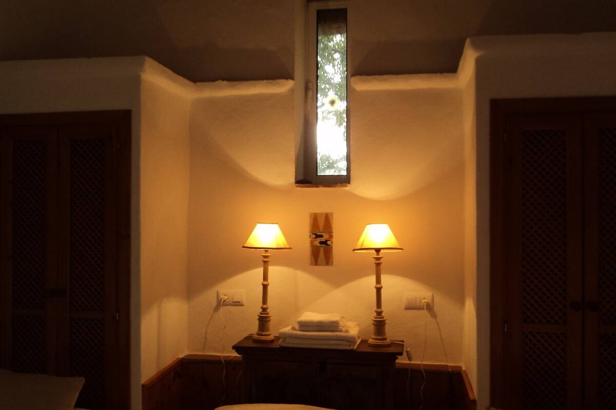 Une pièce faiblement éclairée, avec deux lampes de chevet, une petite fenêtre et une tête de lit en bois.