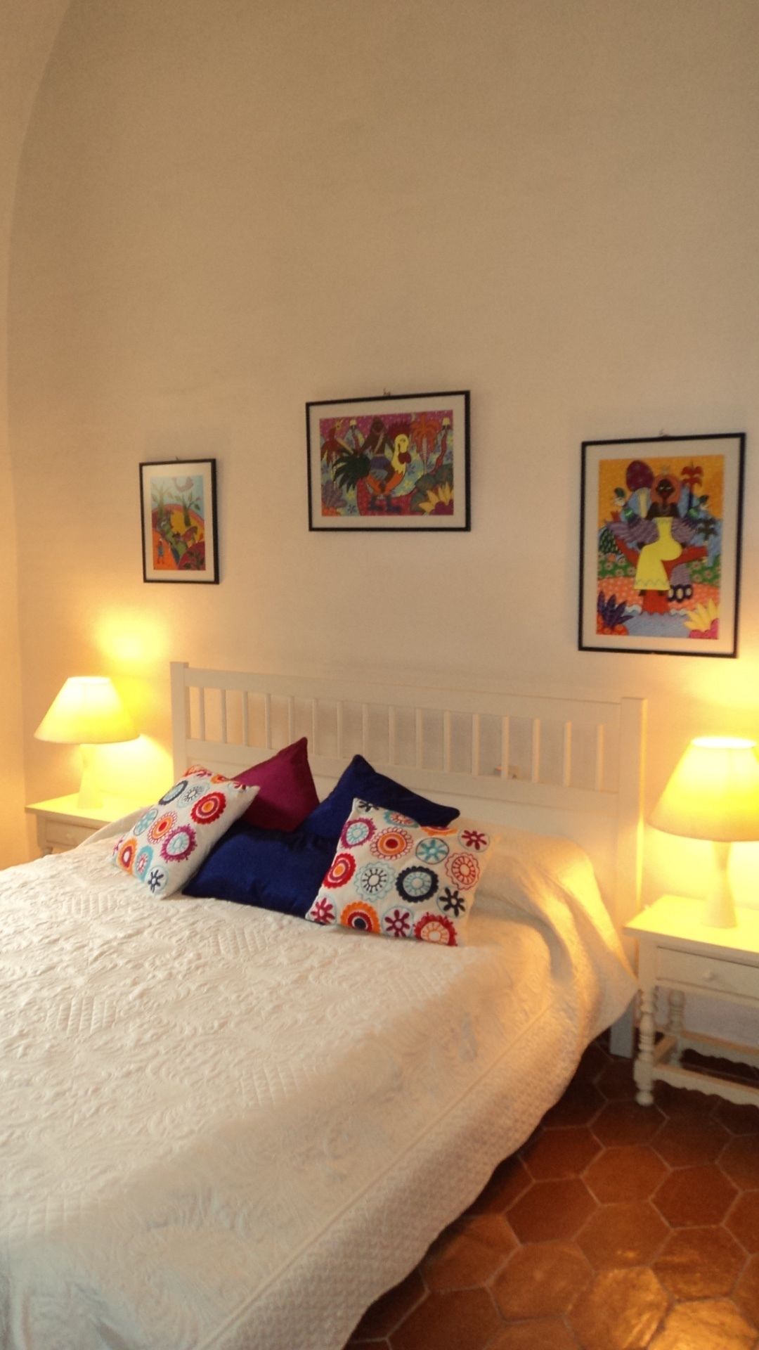 Een bed met een wit hoofdeinde, kleurrijke kussens en drie ingelijste kunstwerken aan de muur.