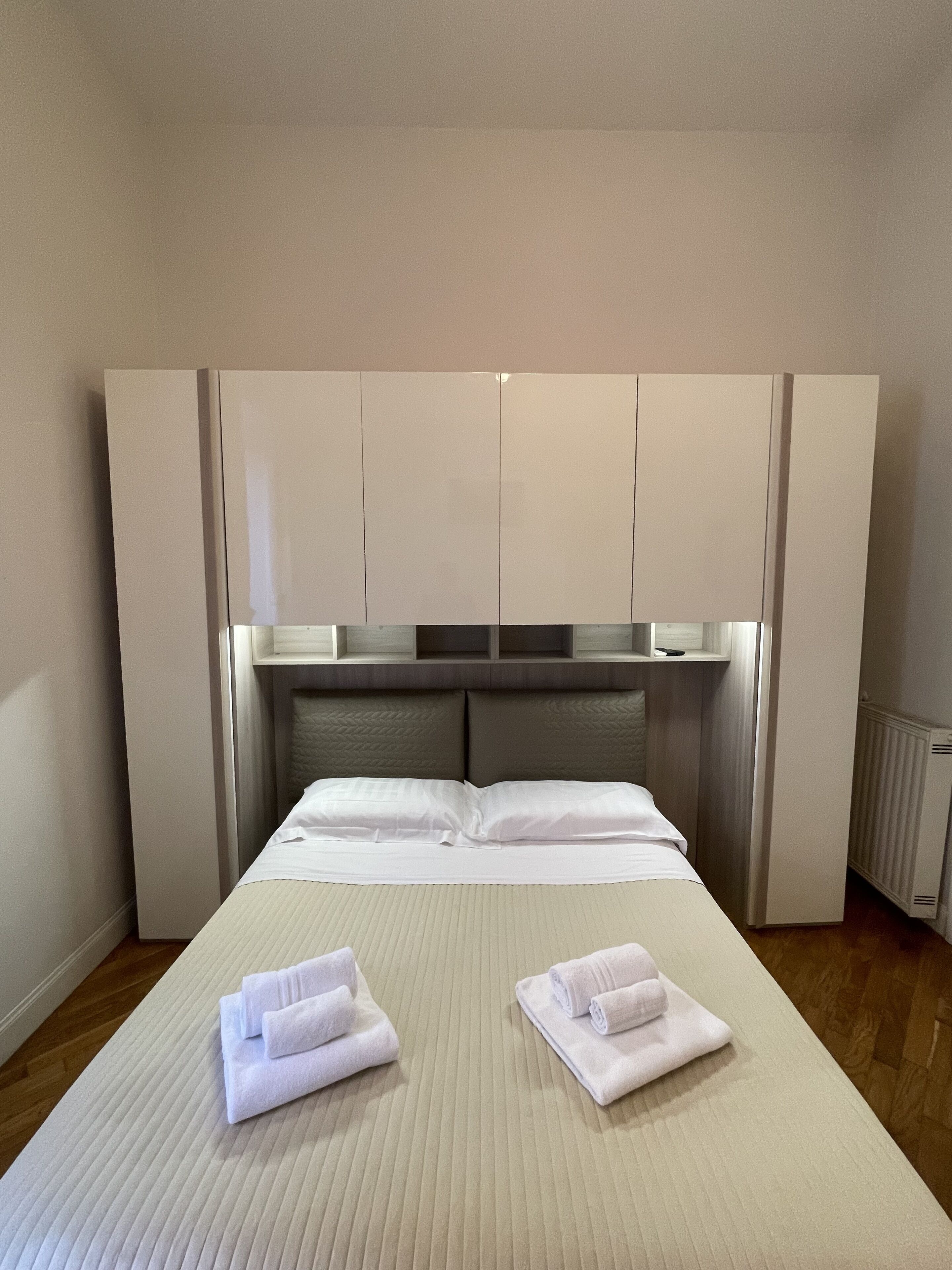 Una cama bien hecha con dos toallas dobladas, un cabecero con compartimentos de almacenamiento y un radiador en la pared.