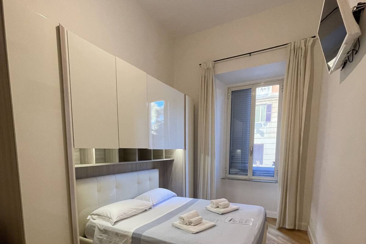 Une chambre à coucher moderne avec un lit, un plancher en bois, une armoire intégrée, une fenêtre avec des rideaux et un téléviseur à écran plat fixé au mur.