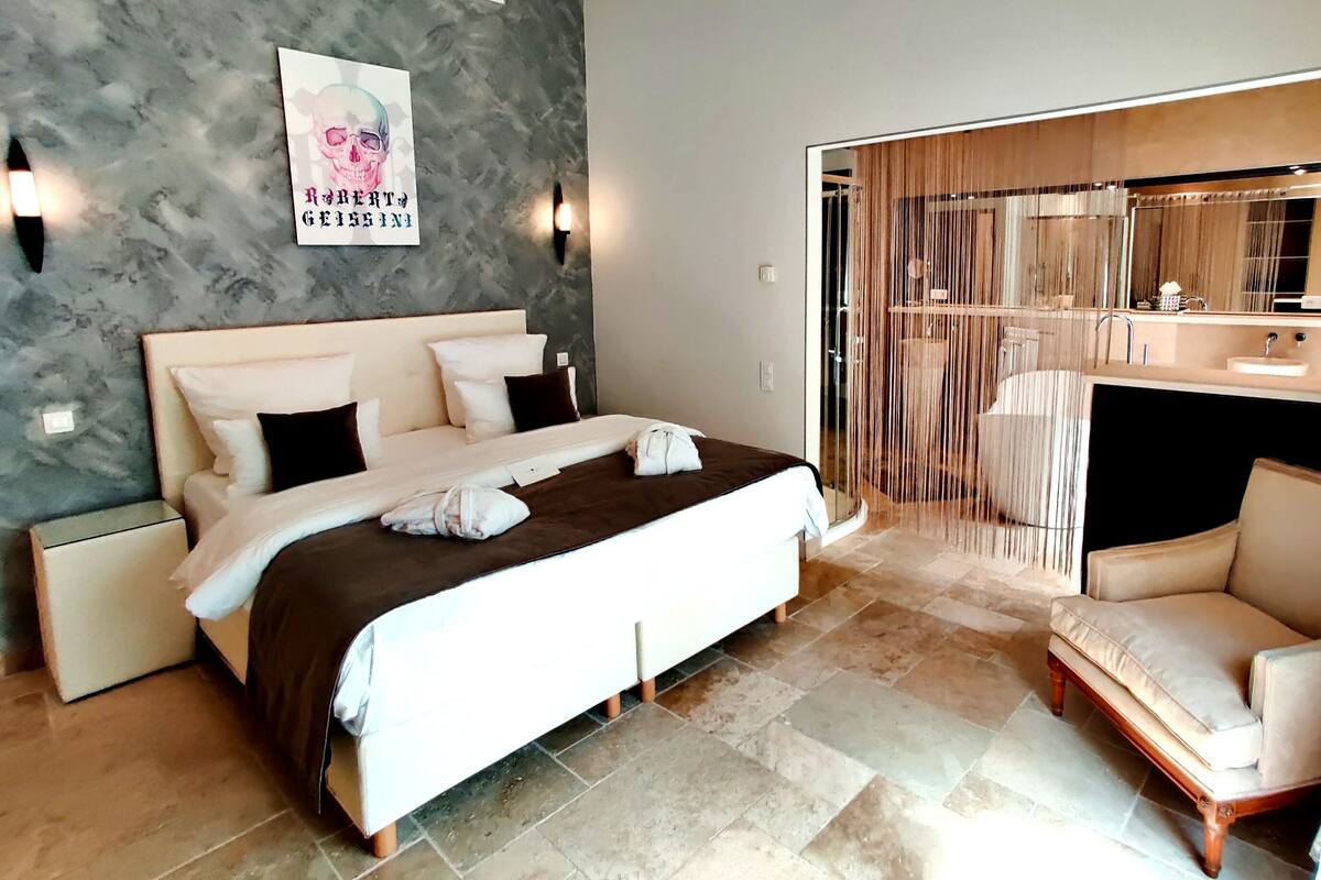 A modern hotel room with a large bed, a bedside table, a chair, a bathroom with a mirror, and a shower area.
