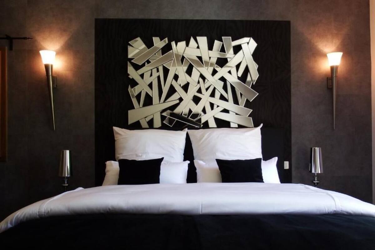 A modern hotel room with a large bed, a geometric wall art piece above the bed, and two wall-mounted lamps on either side.