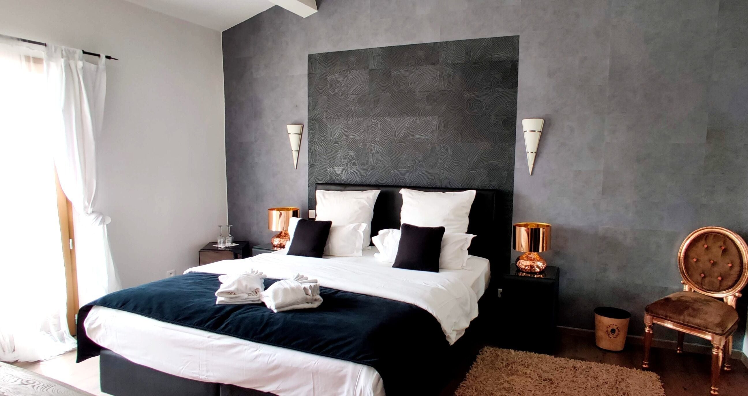 Une chambre à coucher moderne avec un grand lit, une tête de lit sombre, des lampes de chevet et un fauteuil classique.