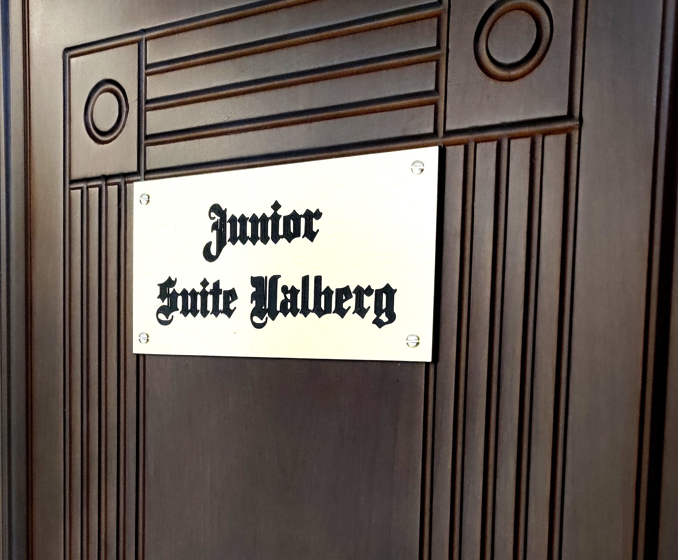 A sign with the text "Junior Suite Halberg" mounted on a wooden panel with vertical lines and decorative elements.