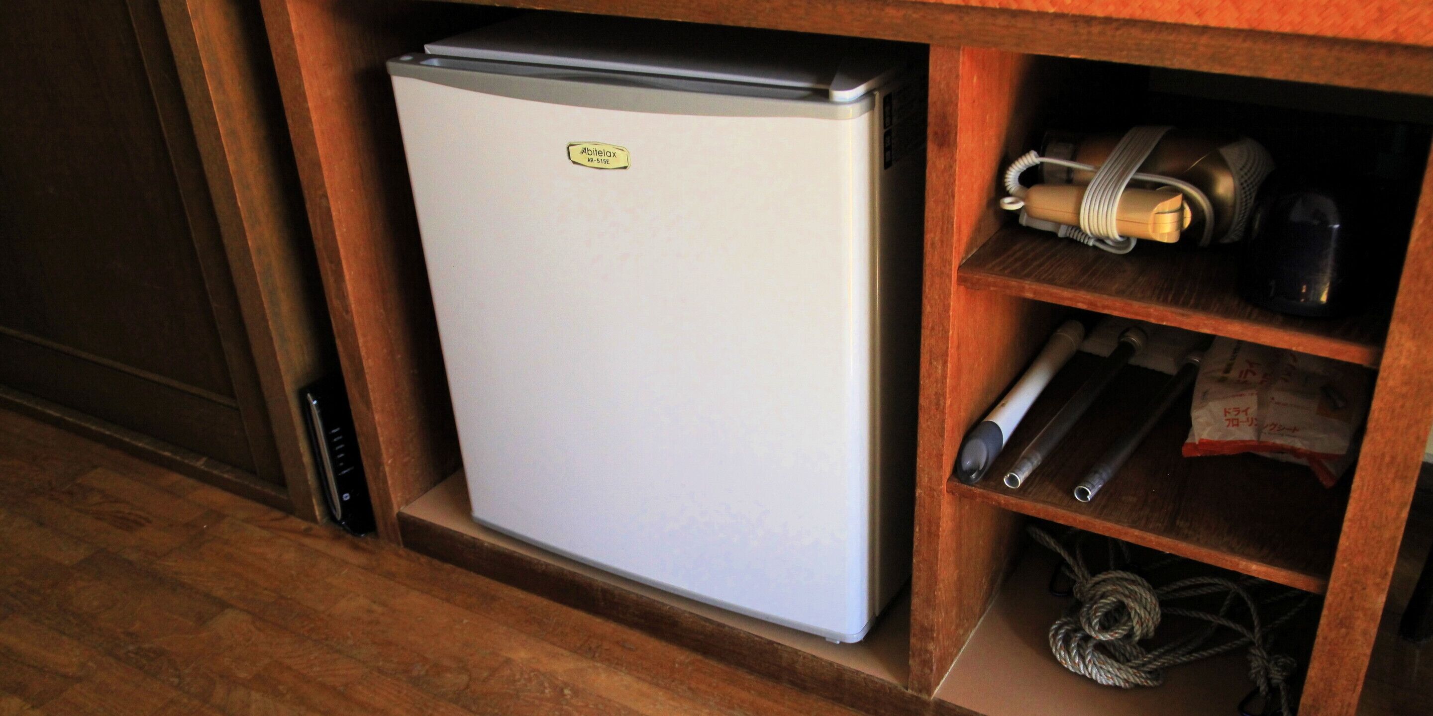 A compact refrigerator in a wooden cabinet with a small shelf containing a bag of snacks and a coiled cable.