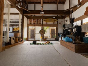 A spacious room with a high ceiling, wooden beams, a large window, and a variety of items including a blue plush toy, a drum, and a guitar. - Holistic retreat coco (Ishigaki)