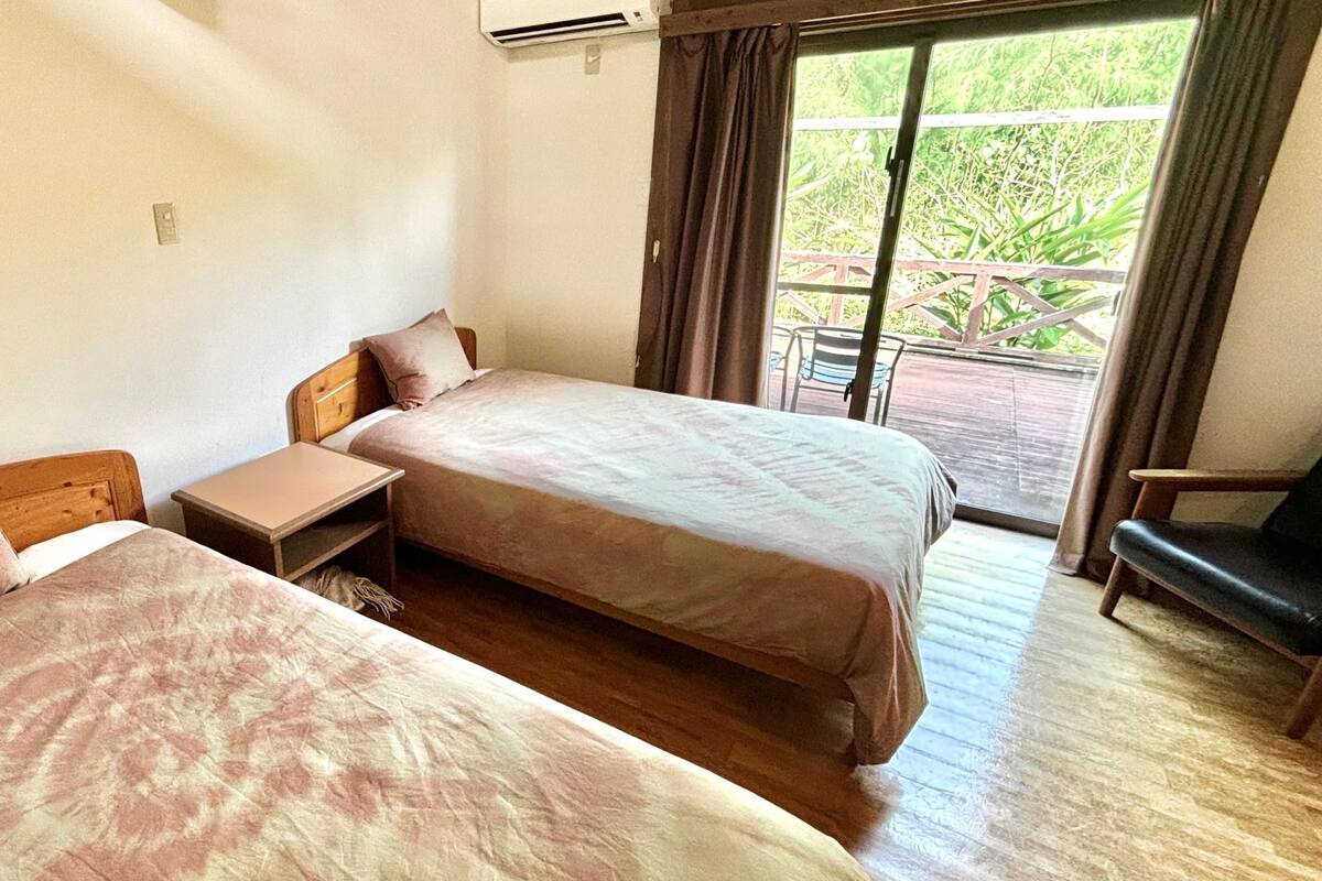 A room with two beds, a small table, and a chair. There is a window with curtains and a view of greenery outside.