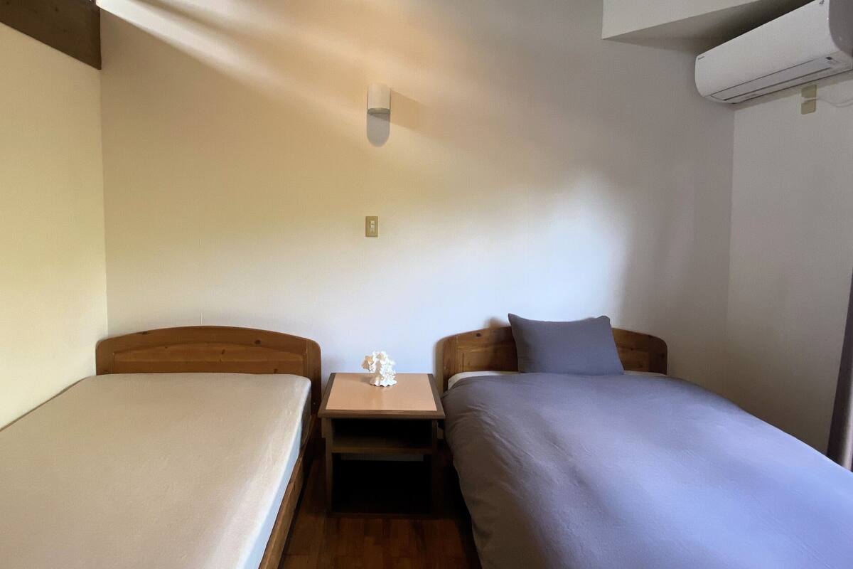 A small, single-room accommodation with two single beds, a nightstand, a wall-mounted air conditioner, and a window with curtains.