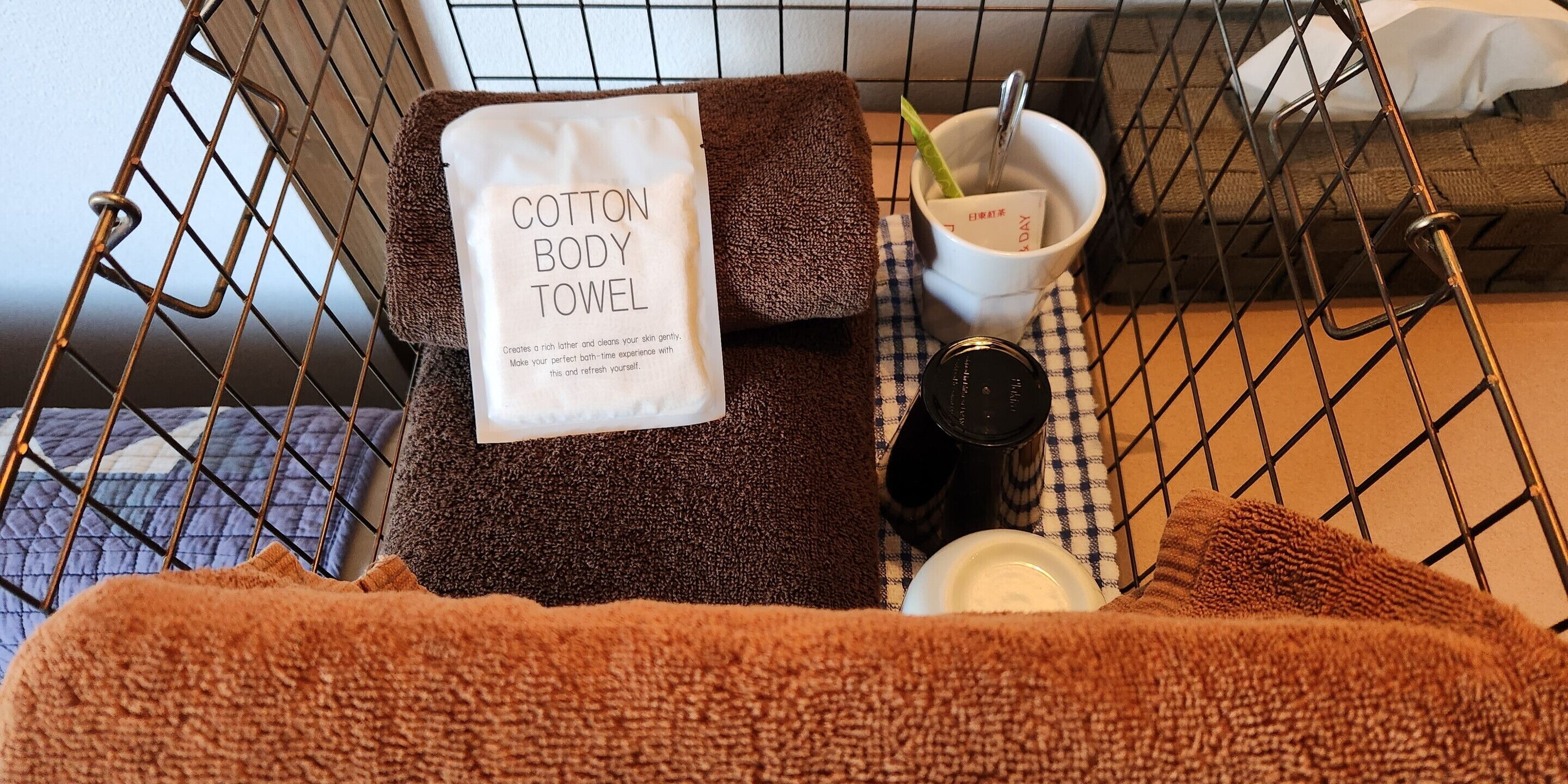 A metal-framed bed with a brown towel, a white cotton body towel, a cup with a spoon, and a bottle on a checkered cloth.