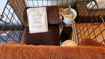 A metal-framed bed with a brown towel, a white cotton body towel, a cup with a spoon, and a bottle on a checkered cloth.